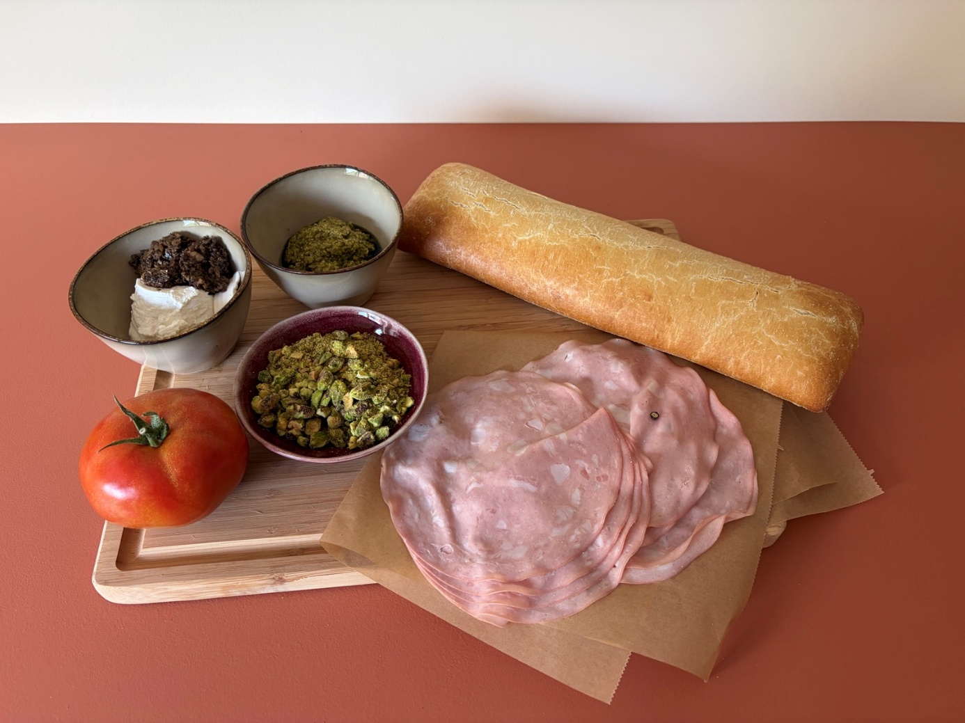 Ingrediënten op een snijplank van links naar rechts: een bakje met ricotta en truffeltapenade, één tomaat, groene pesto, gemalen pistachenootjes, mortadella en één baguette