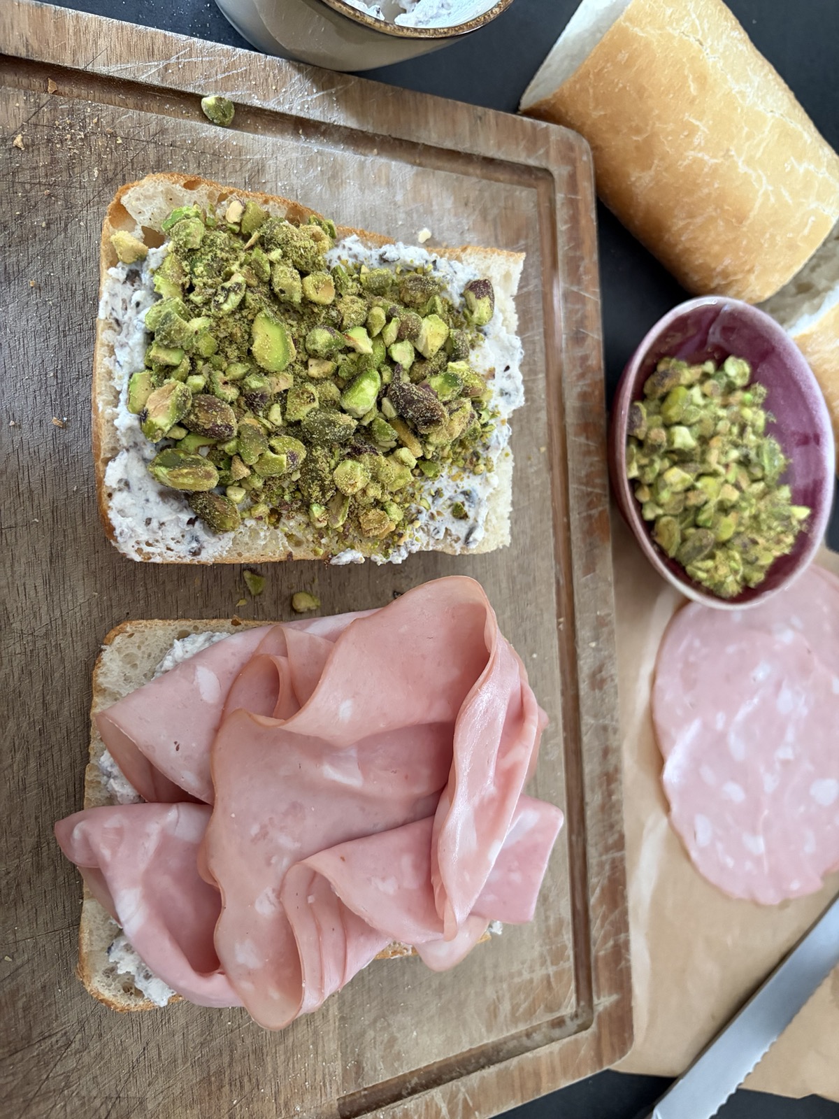 Broodje belegd met tomaat, pesto, de ricotta-truffelmix, pistachenootjes en mortadella op een snijplank, met alle ingrediënten op de achtergrond