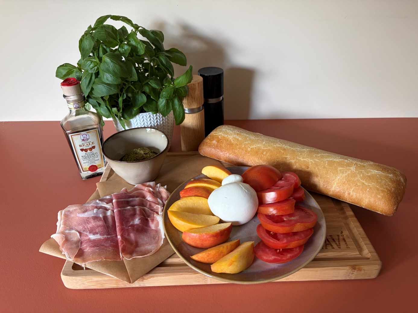 Ingrediënten op een snijplank van links naar rechts: balsamico, verse basilicum, groene pesto, parmaham, zout, peper, een rond bord met nectarinepartjes, burrata, gesneden tomaat en een baguette