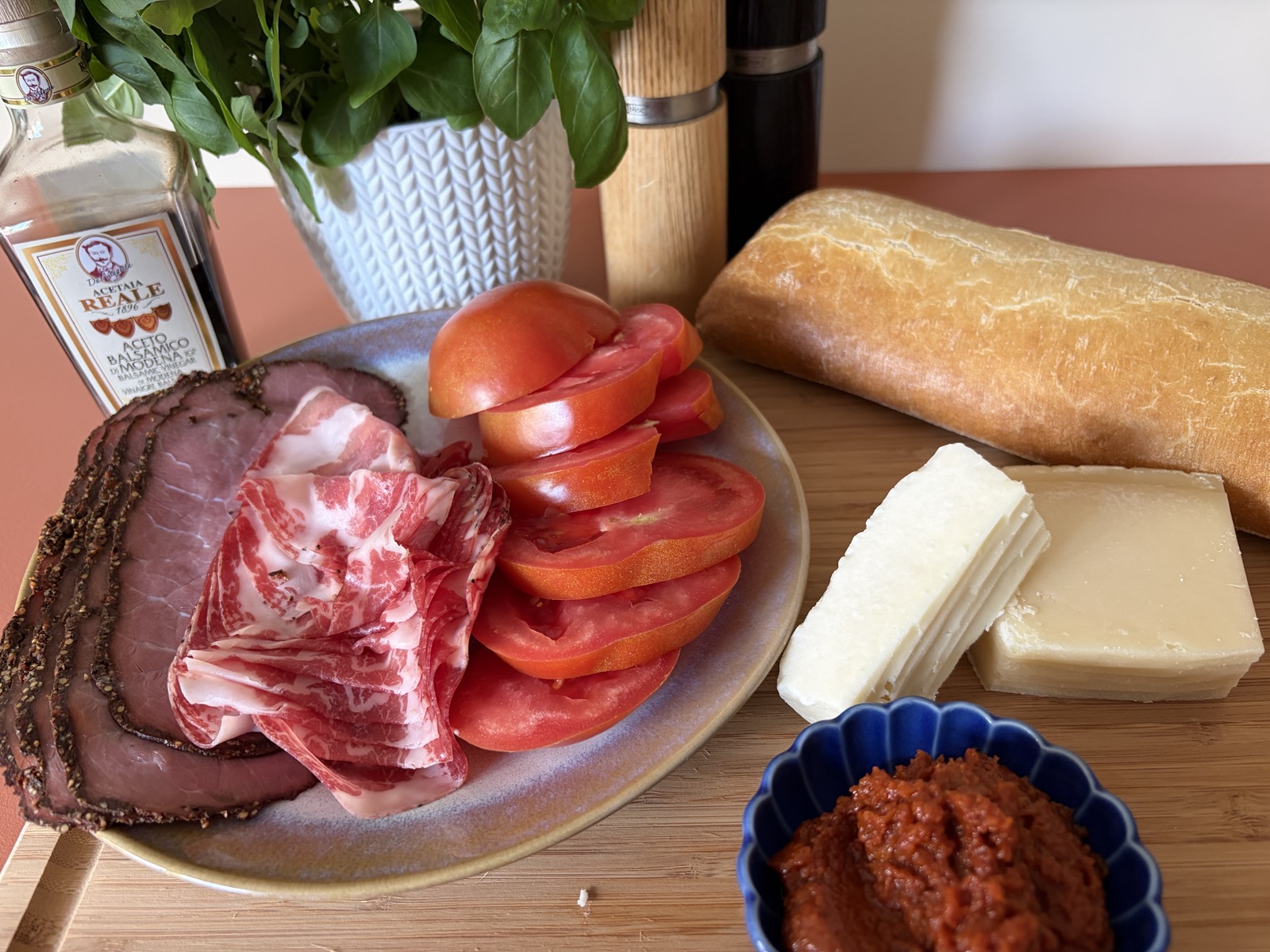 Ingrediënten op een snijplank van links naar rechts: balsamico, verse basilicum, pastrami, coppa di parma, plakken tomaat, zout en peper, pecorino en Parmezaanse kaas, een blauw schaaltje met zongedroogde-tomatentapenade en een baguette