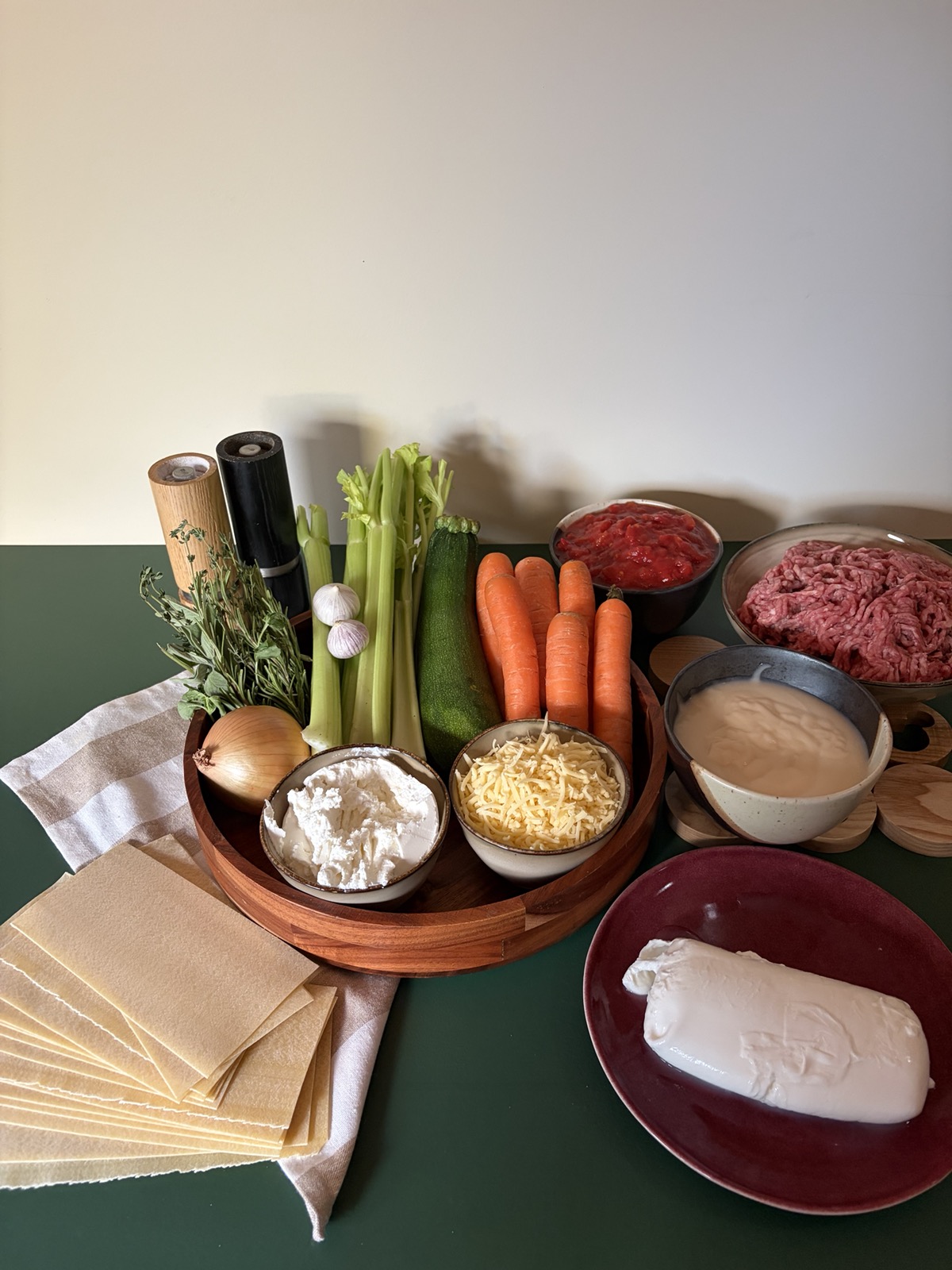 Alle ingrediënten van links naar rechts: lasagnevellen, peper- en zoutmolen, verse Italiaanse kruiden, een witte ui, twee bollen knoflook, bleekselderij, courgette, winterpenen, een kom ricotta, geraspte jonge kaas, sugocasa, gehakt en een mozzarella