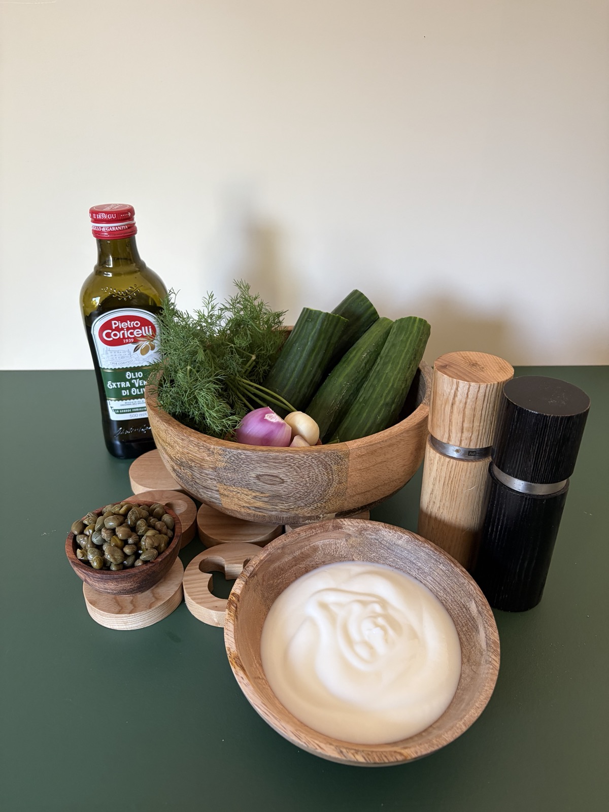 Alle ingrediënten van links naar rechts: extra vierge olijfolie, een schaaltje kappertjes, verse dille, een sjalot, twee teentjes knoflook, komkommers, yoghurt en een peper- en zoutmolen