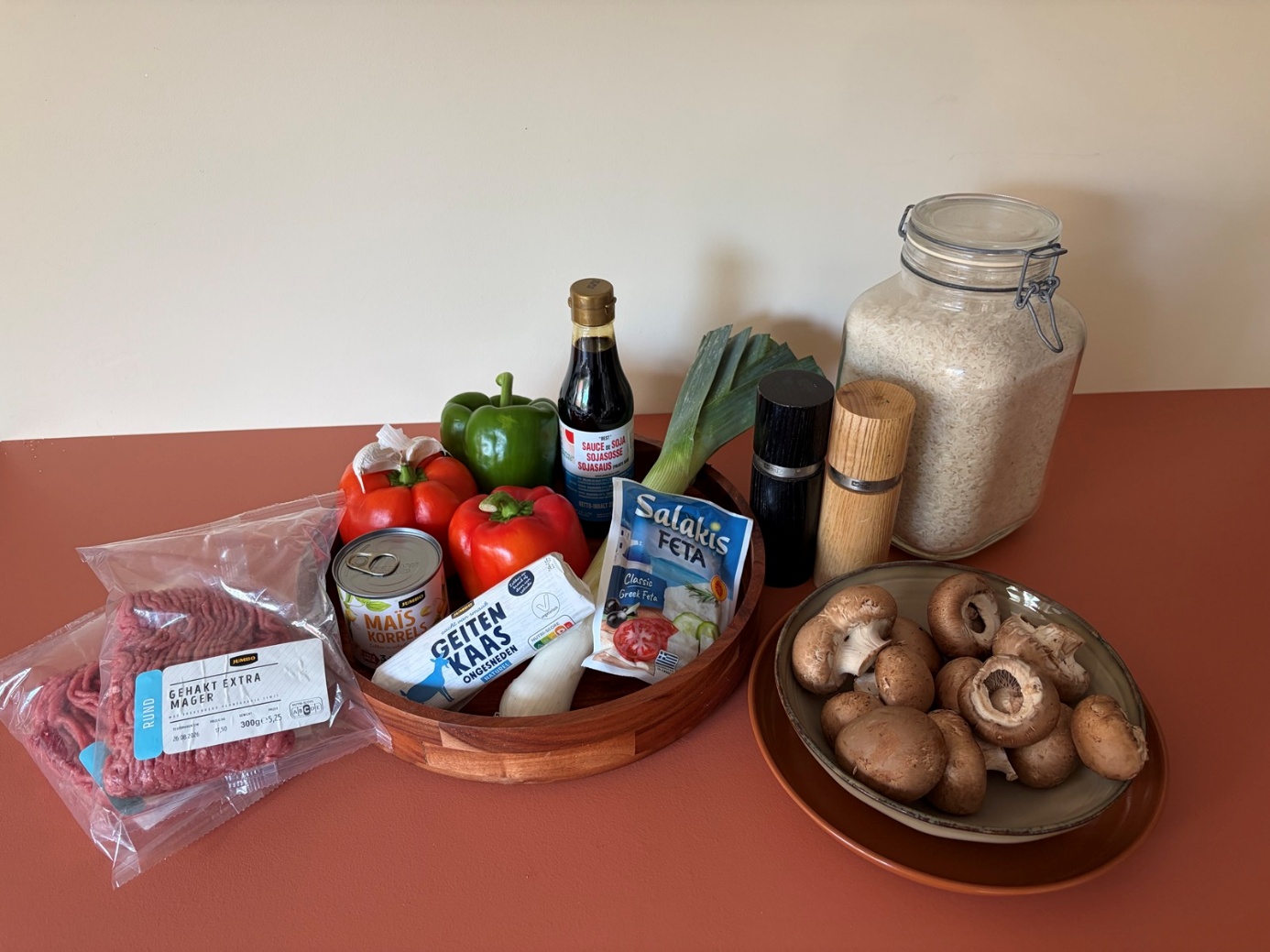 Alle ingrediënten van links naar rechts: extra mager rundergehakt, drie paprika's, donkere sojasaus, een klein blikje maiskorrels, een rolletje geitenkaas, één prei, een pak feta, zwarte peper, zout, pandanrijst en kastanjechampignons op een bord