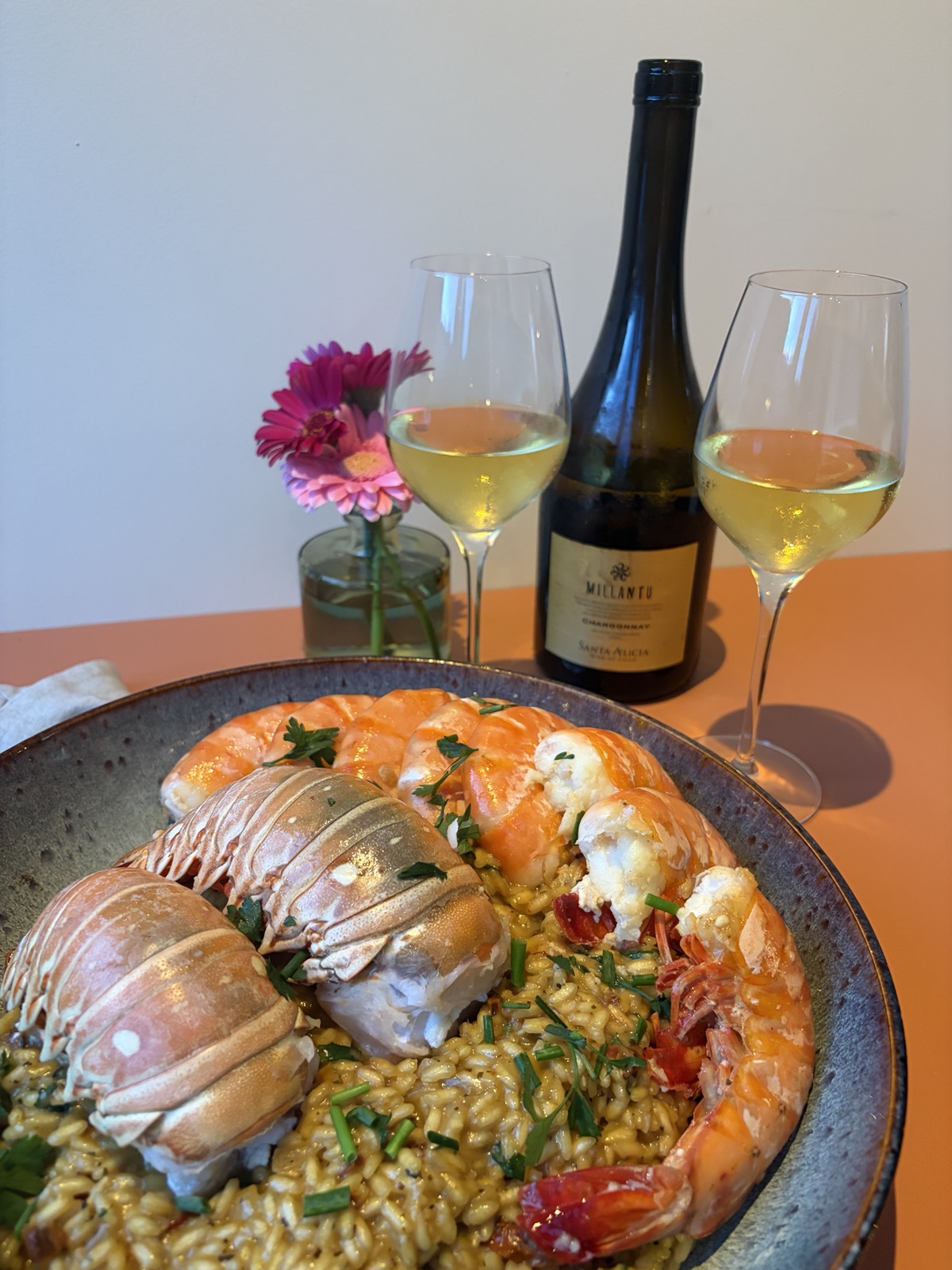 Een grote schaal met de risotto, kreeftenstaarten en garnalen, met bloemen en twee glazen witte wijn en de fles erbij