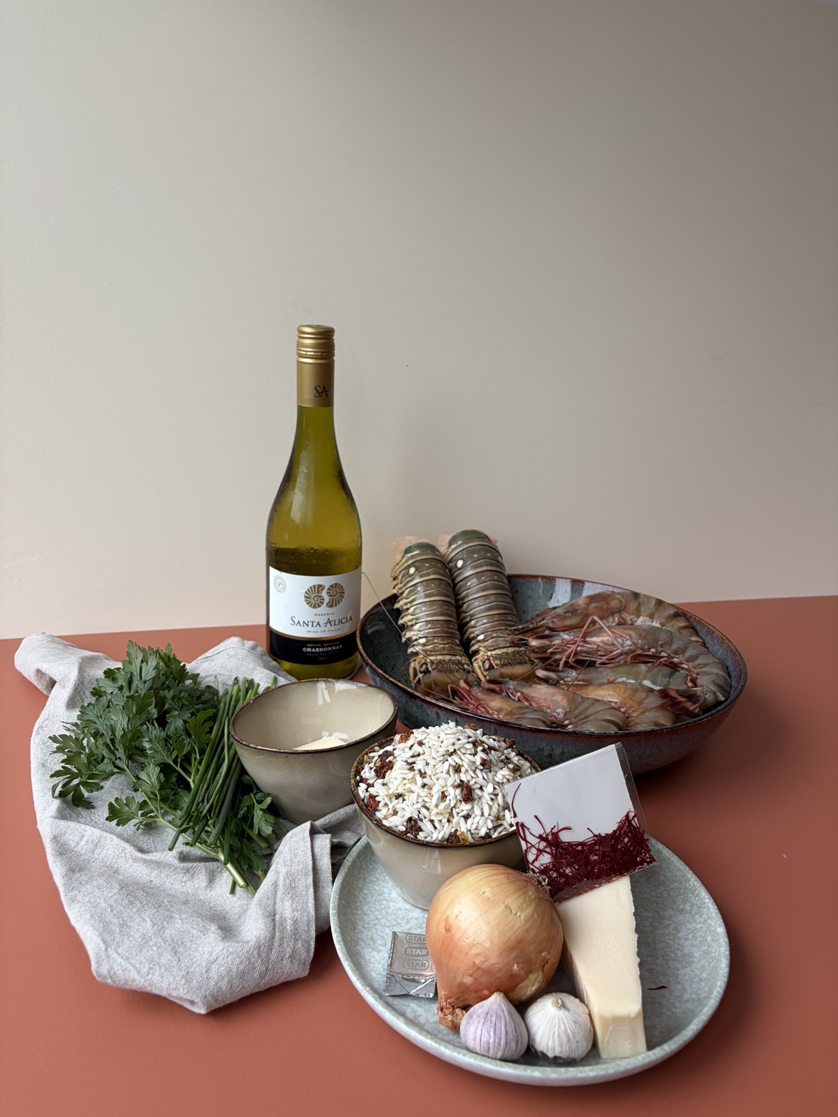 Alle ingrediënten: verse peterselie en bieslook op een doek, een bakje roomboter, een fles witte chardonnay, risottorijst met zongedroogde tomaat, een risottobouillonblokje, een witte ui, twee bollen knoflook, saffraan, Parmezaanse kaas en een schaal met twee kreeftenstaarten en black tiger garnalen