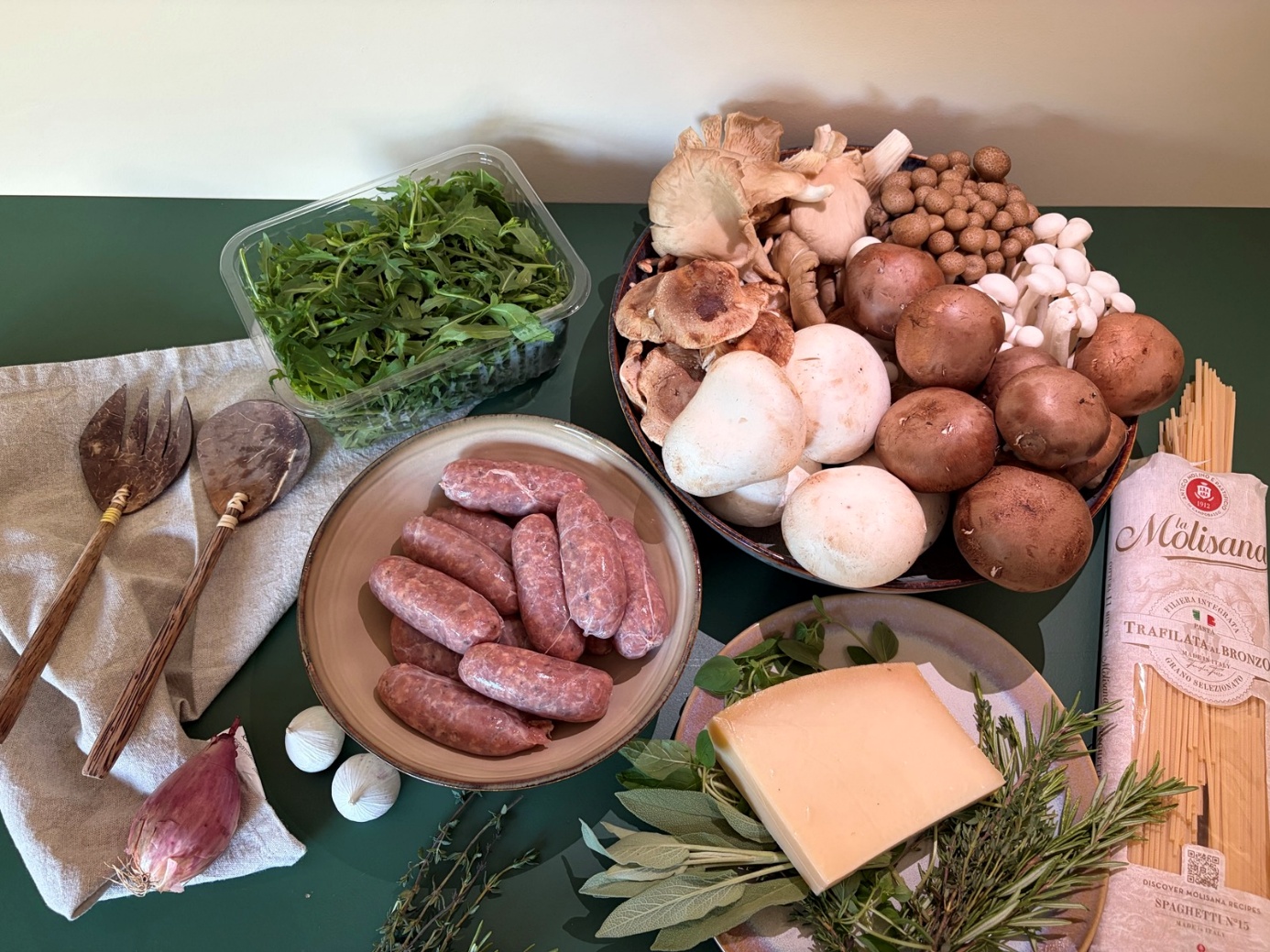 De ingrediënten van links naar rechts met een opscheplepel en -vork: sjalot, twee grote bollen knoflook, rucola, worstjes op een rond diep bord, een overzicht van alle paddenstoelen, een bord met de kruiden en Parmezaanse kaas, en spaghetti